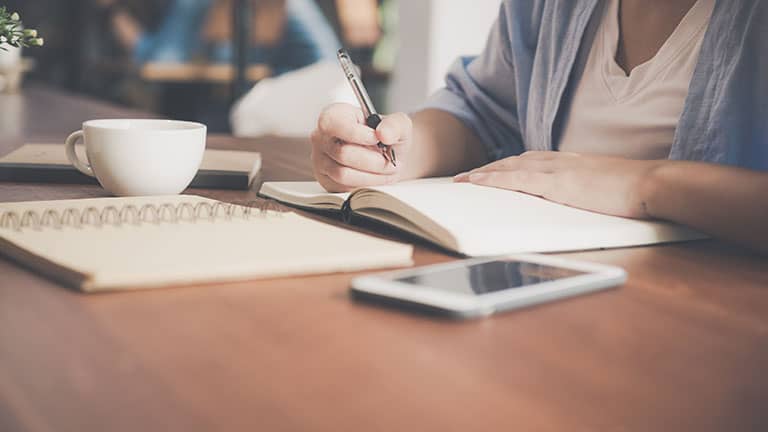 Woman writing on a notebook beside teacup and tablet